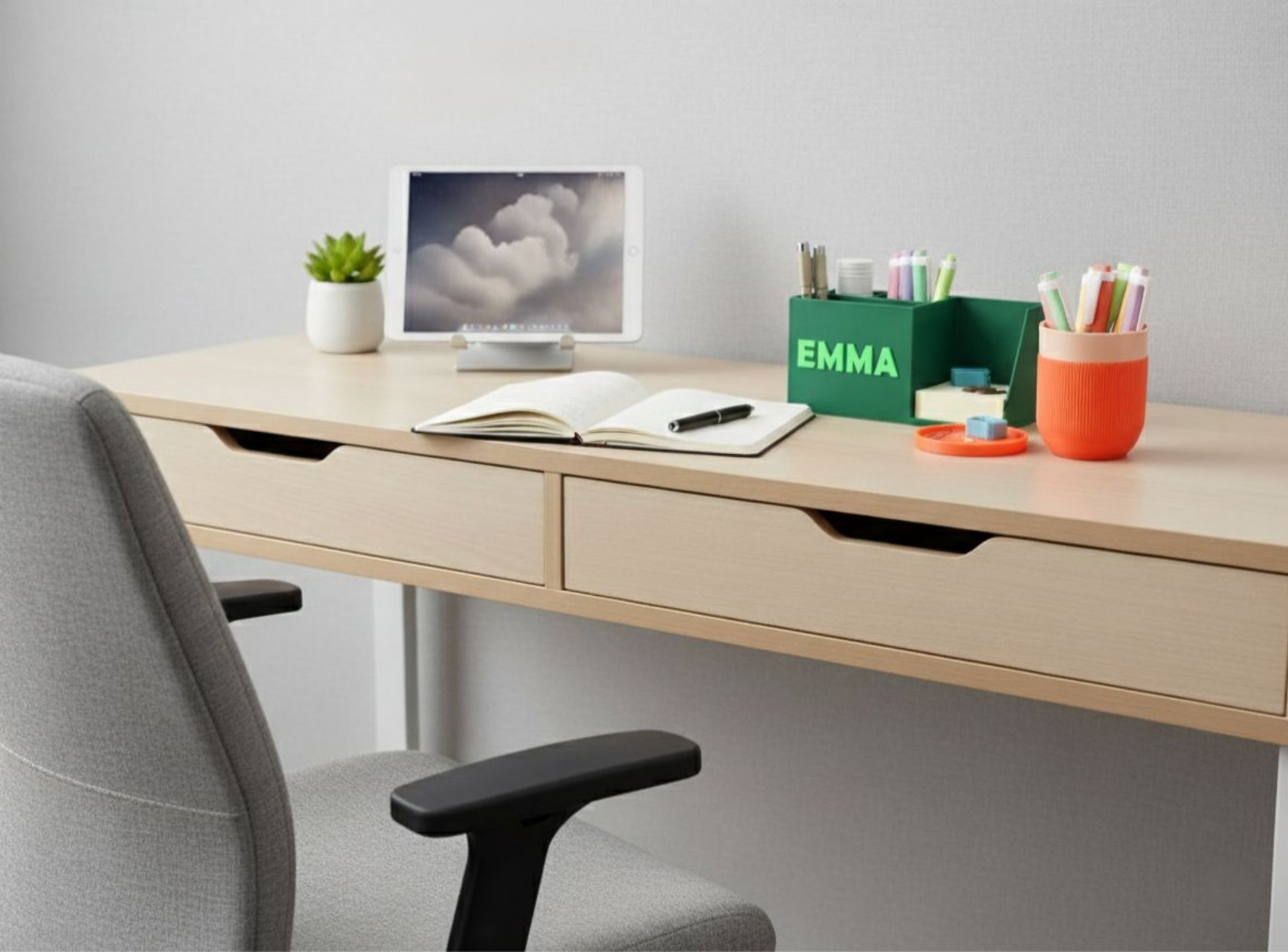 Modern office desk with a chair, computer monitor, notebook, and stationery items.